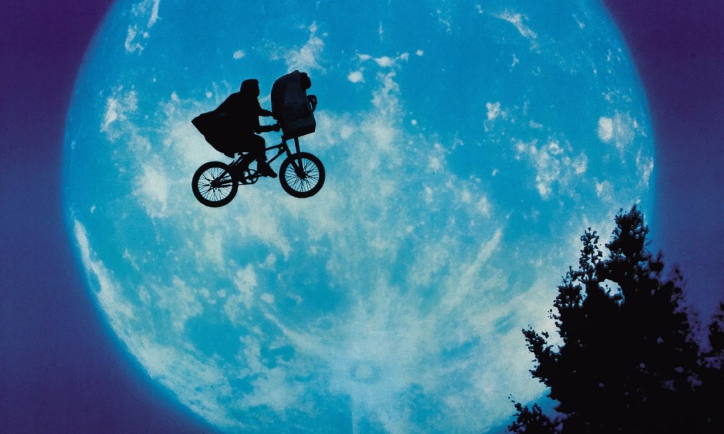 A boy rides his bike in the sky with E.T., an alien in the front basket. The picture shows them in from of a giant moon