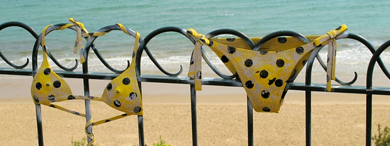 A yellow polka dot bikini rests on a metal fence near the beach.