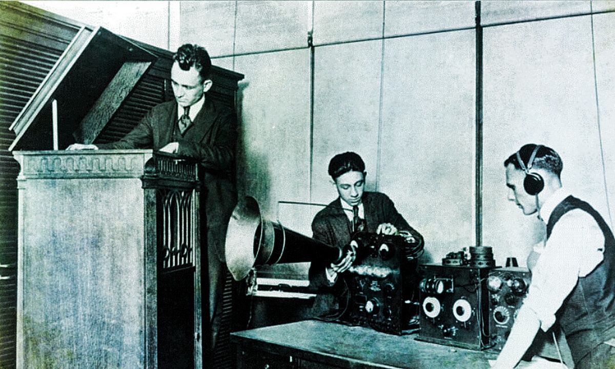 Three men work in the recording room of an early radio station.