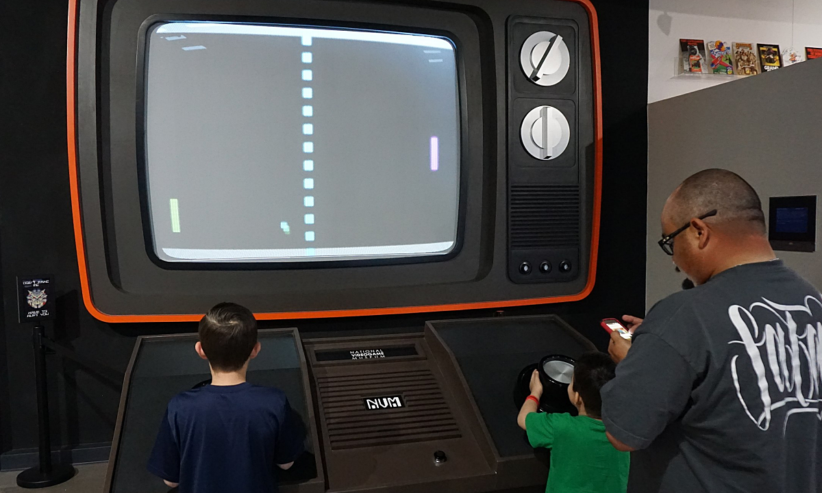 Two boys play a game of Pong using a larger than life video game console