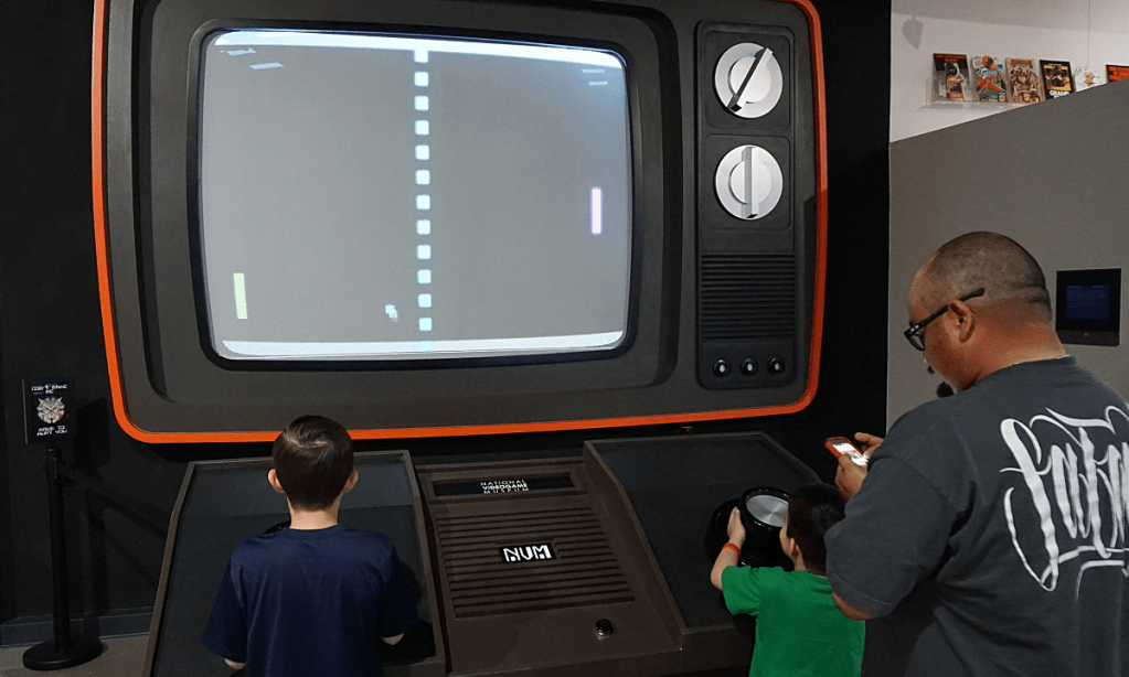 Two boys play a game of Pong using a larger than life video game console