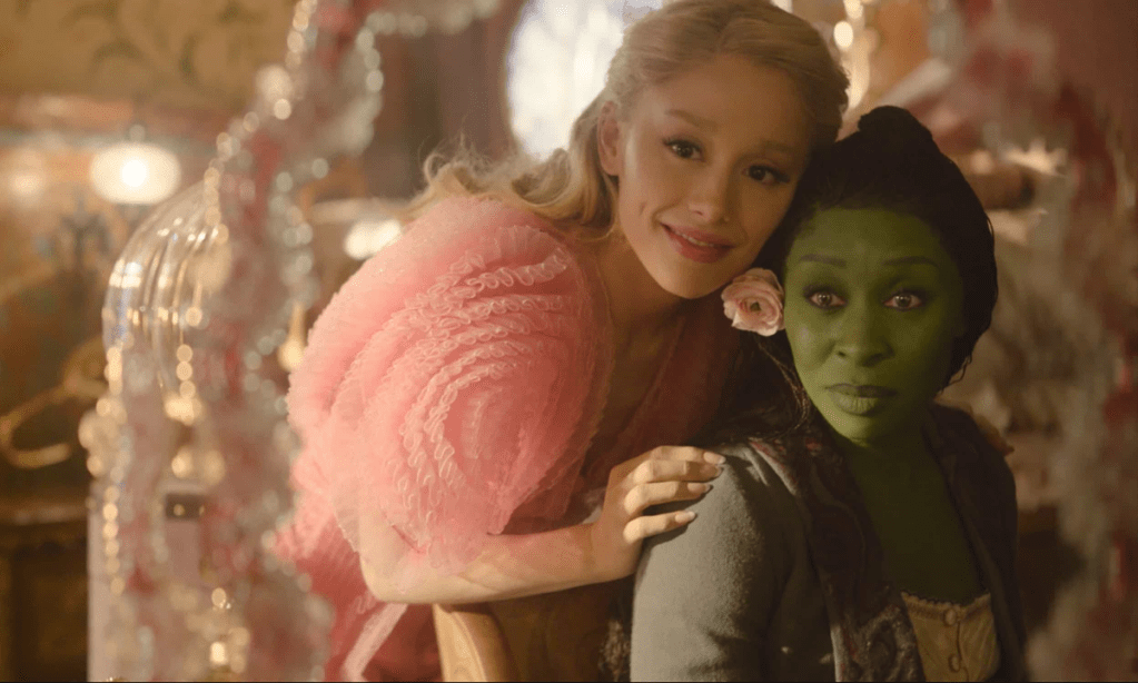 Pink-clothed Galinda leans over green-skinned Elphaba looking forward (From Wicked)
